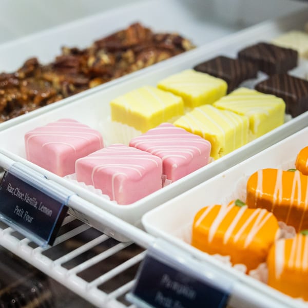 Trays of colorful petit fours, including pink, yellow, and orange, displayed in a deli case.
