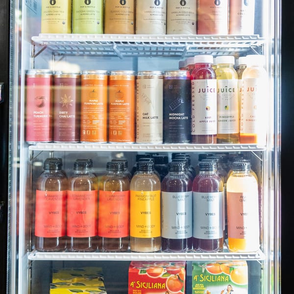 A deli cooler filled with various bottled and canned beverages, including juices, lattes, and mood drinks.